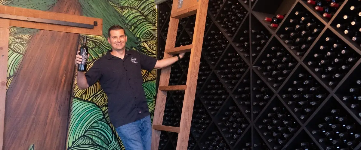 A man in a dark polo shirt stands indoors beside a large wooden crate wall of wine bottles. He holds up a wine bottle in one hand and gestures with the other near a wooden ladder-like frame. Colorful painted wall art with swirling green and orange sw