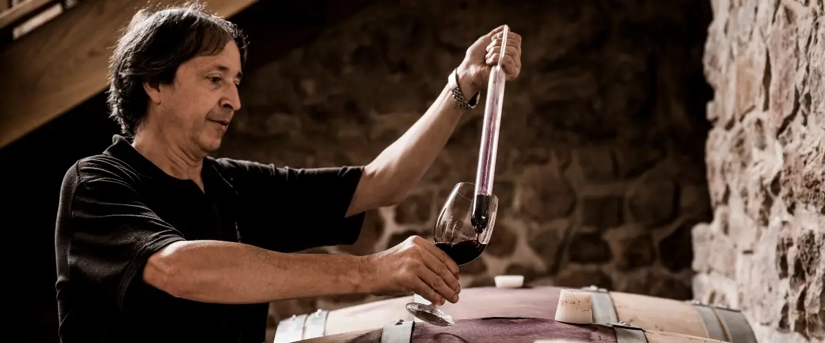 A man in a black shirt pours a dark red liquid from a wine bottle into a glass held on a wooden table. Candles and small items sit on the table, with a stone wall in the background.