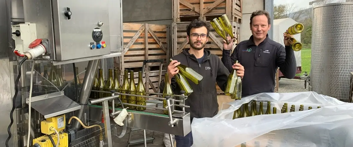 Two men stand beside an industrial wine bottling setup. A machine line with metal parts and a control panel is on the left, while multiple green glass bottles are on the table and trays. The men smile and each holds several capped green wine bottles.