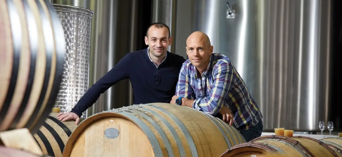 Two men stand behind and rest their hands on large wooden wine barrels in a winery cellar. Stainless steel tanks and a metal-framed barrel rack are visible in the background, with an additional barrel in the foreground.