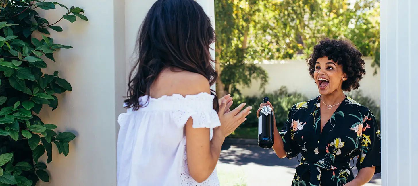 Two women stand in a sunny outdoor setting by a white wall and green foliage. One woman faces away wearing a white off-shoulder dress, gesturing toward the other who is smiling broadly in a dark floral shirt while holding a glass bottle with a drink.